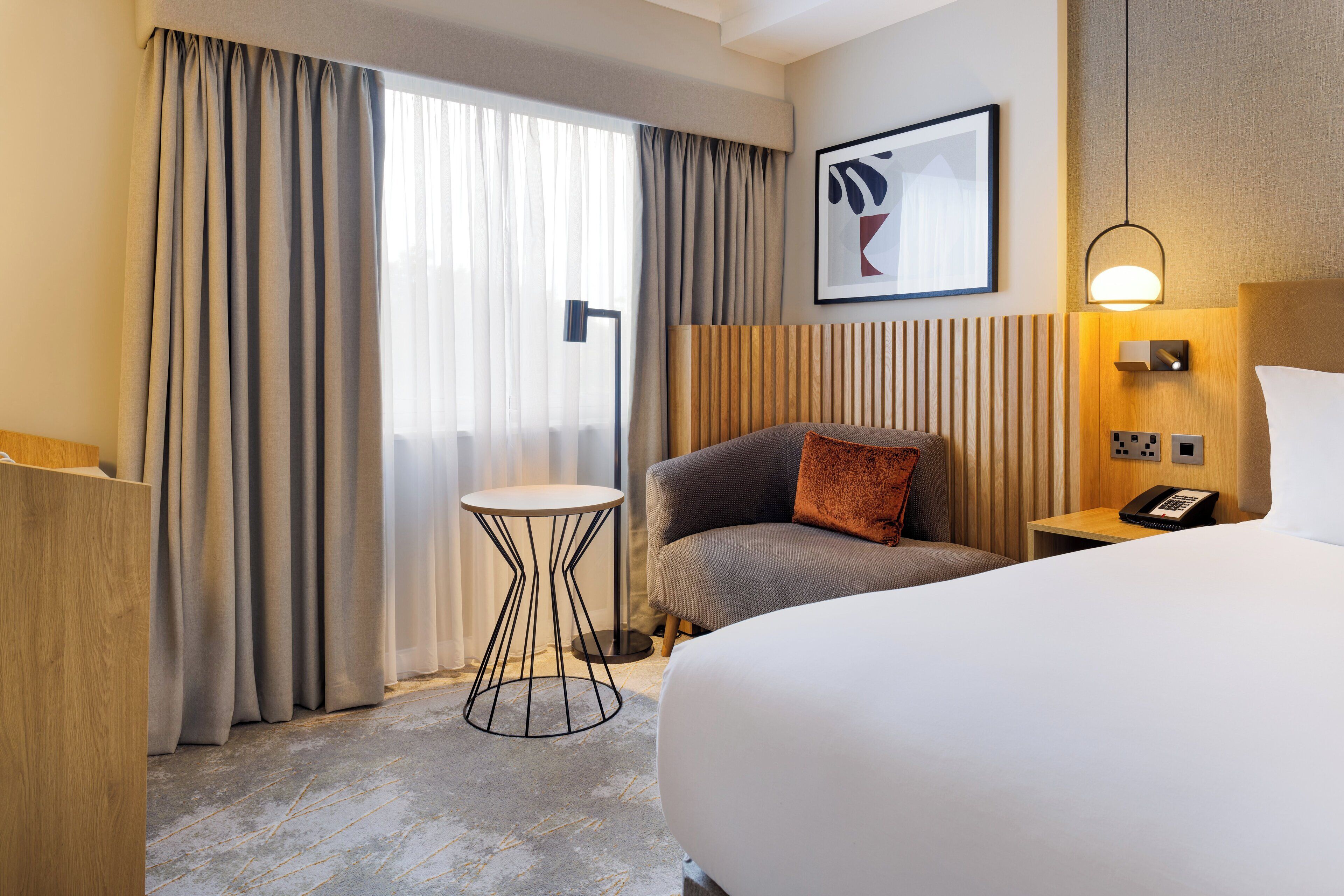 Une chambre d’hôtel avec un grand lit, une petite table ronde, une chaise, un bureau avec un téléphone et un tableau encadré au mur.