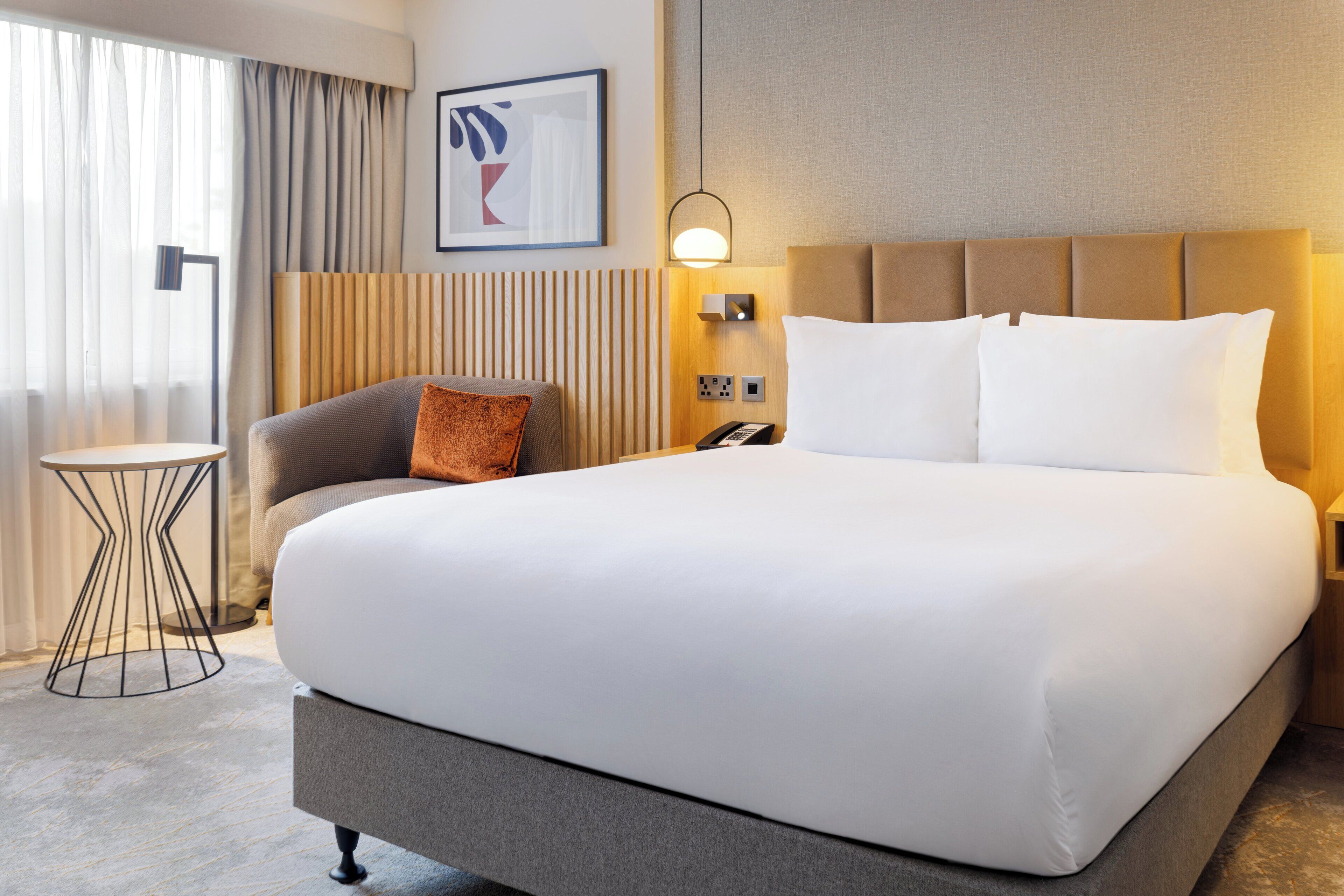 Ein Hotelzimmer mit einem großen Bett, einem Nachttisch, einer Lampe, einem gerahmten Kunstwerk an der Wand und einem Fenster mit Vorhängen.