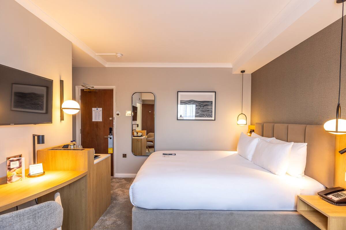 A hotel room with a large bed, bedside tables, a desk, a mirror, and artwork on the walls.