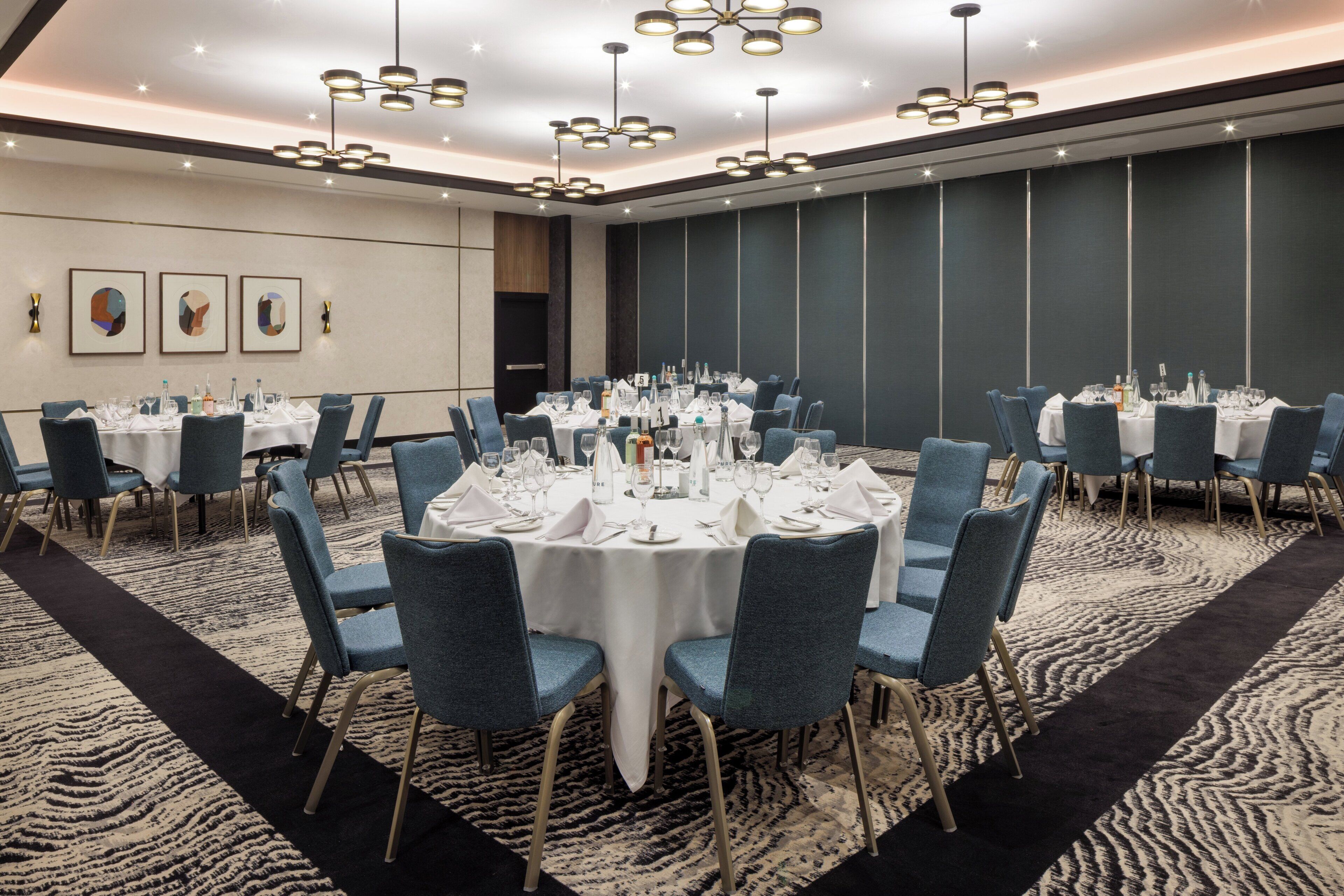 A conference room with round tables, blue chairs, and white tablecloths, featuring modern lighting and a patterned carpet.
