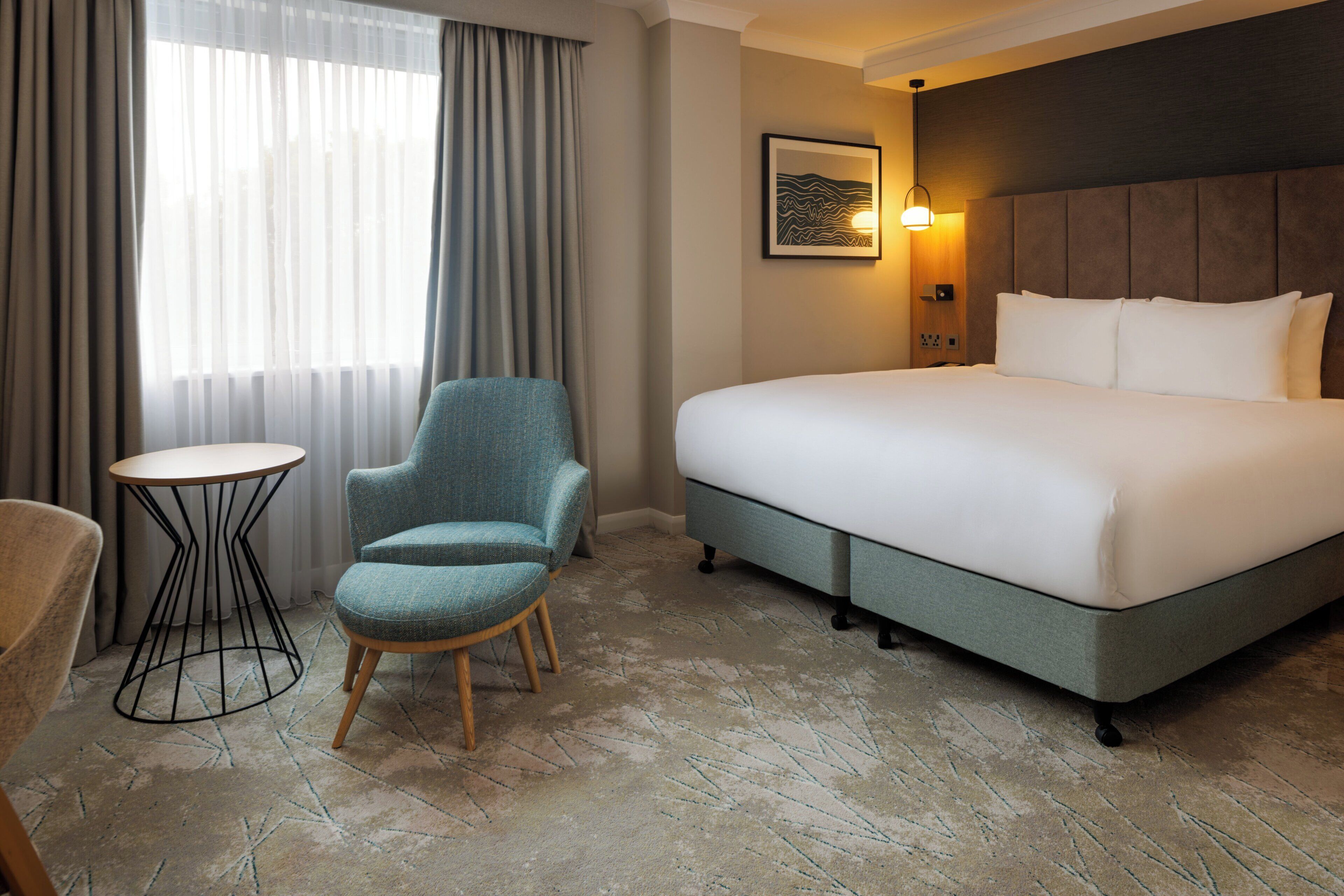Une chambre d’hôtel avec un grand lit, une petite table ronde, un fauteuil bleu et un luminaire fixé au mur.