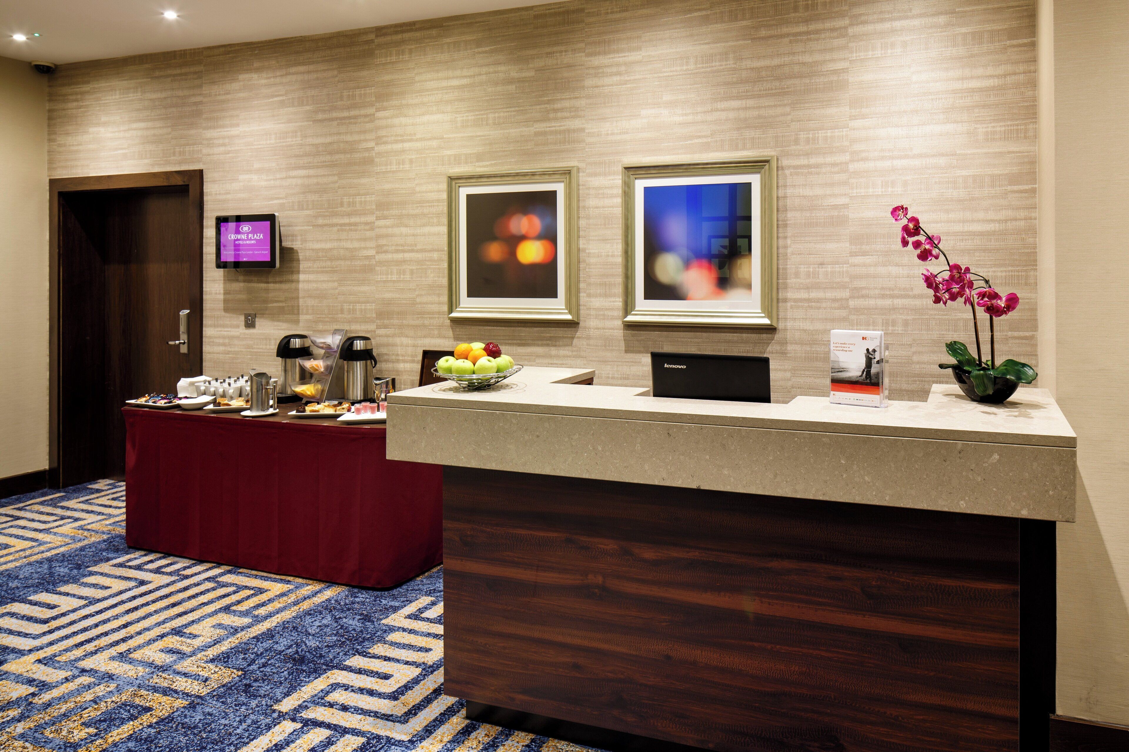 A hotel reception area with a reception desk, a digital screen, framed artwork, and a fruit bowl.