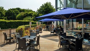 Un patio exterior con mesas y sillas, cubierto por sombrillas azules, y un cerramiento de vidrio con vista al jardín.
