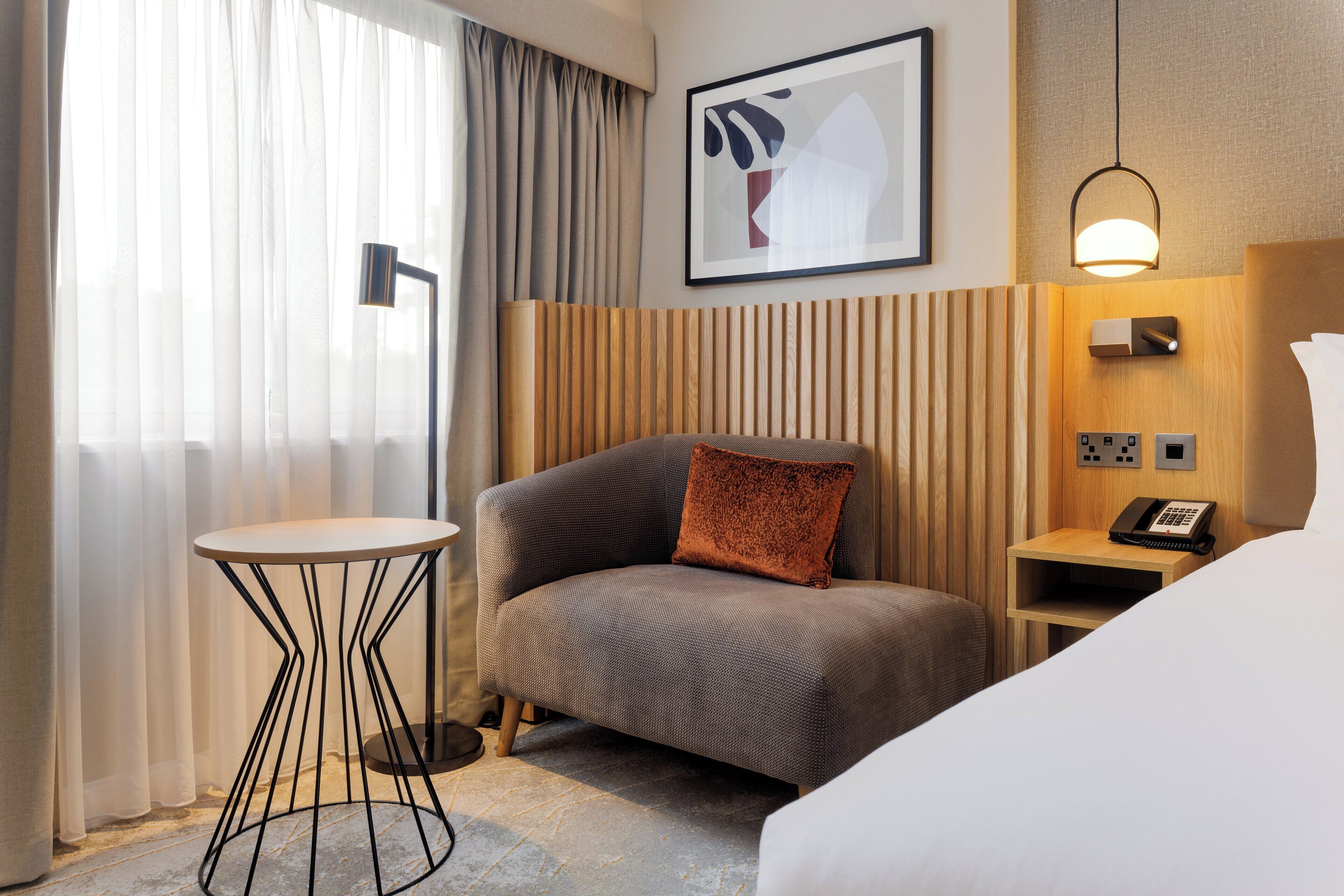 A hotel room with a bed, a small table, a chair, a bedside table with a phone, and a wall-mounted light fixture.