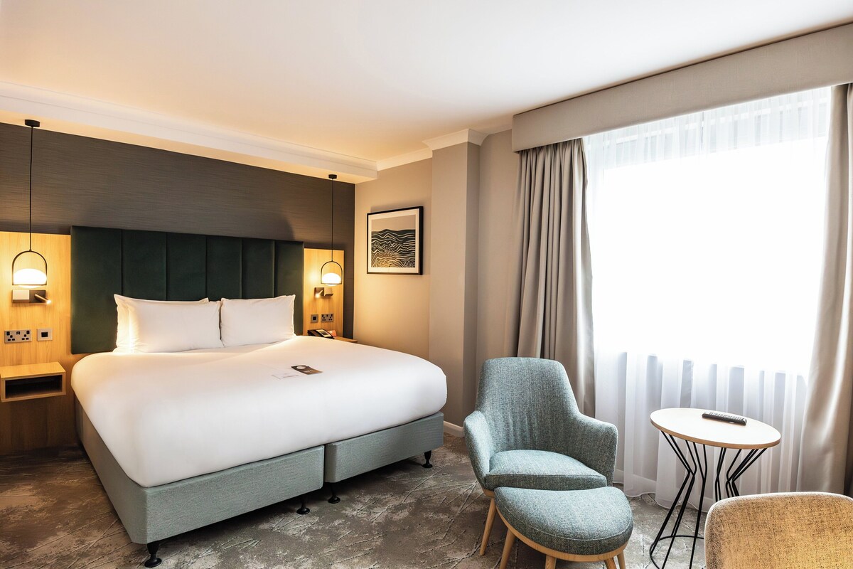 Ein Hotelzimmer mit einem großen Bett, einem kleinen runden Tisch, zwei Stühlen und Blick ins Freie durch ein Fenster mit Vorhängen.