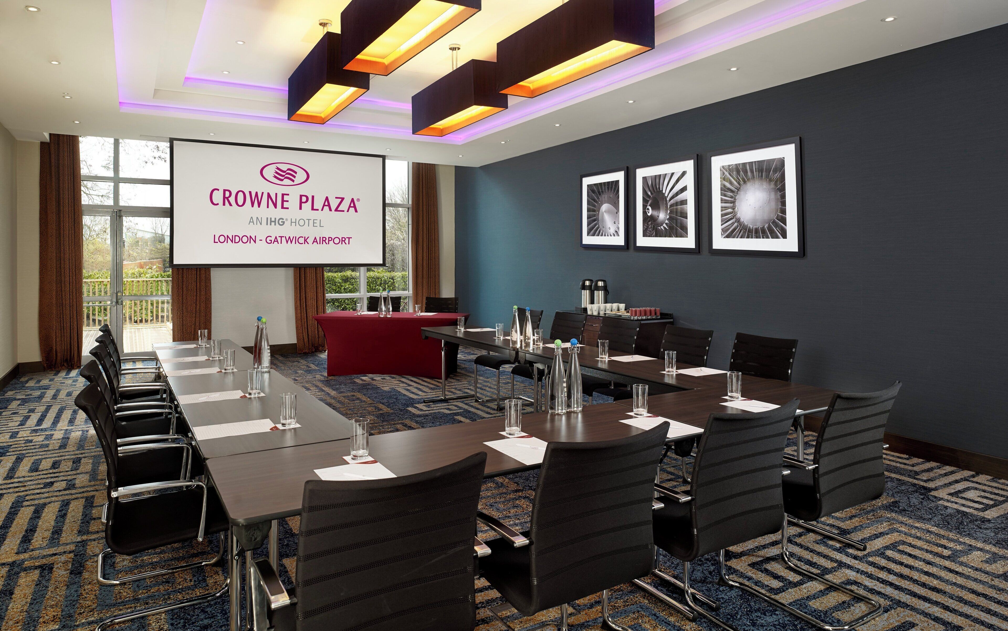Un salón de conferencias en Crowne Plaza, Londres - Aeropuerto de Gatwick, con una pantalla grande que muestra el nombre del hotel, una larga mesa con sillas y obras de arte enmarcadas en la pared.