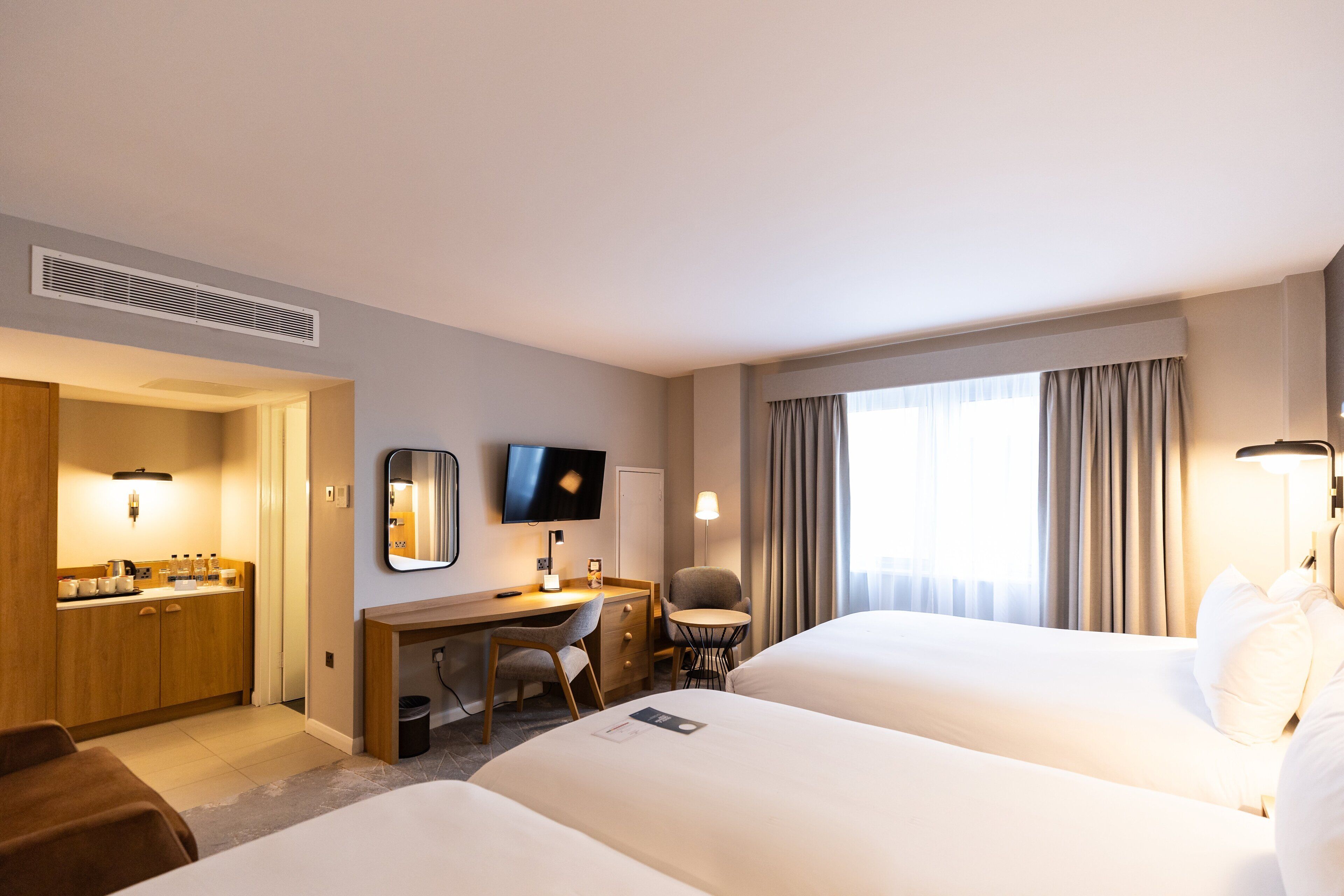Habitación de hotel con dos camas, un escritorio con silla, televisor y baño con lavamanos y espejo.