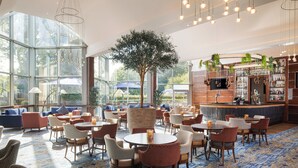 Un espacio interior amplio con mesas y sillas redondas, un gran árbol y una zona de bar con estantes llenos de botellas.