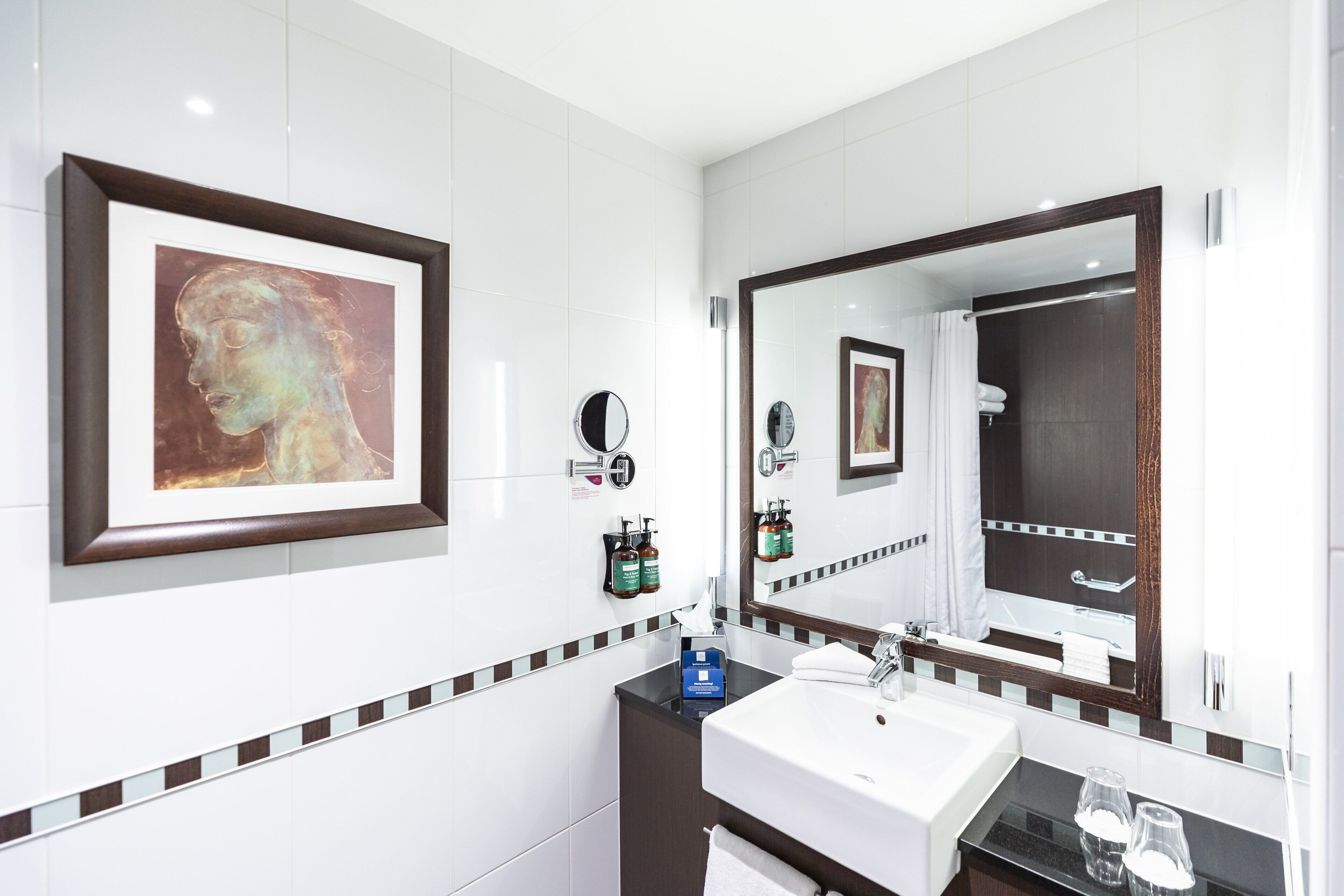 A modern bathroom with a large mirror, a framed artwork, a sink, and toiletries.