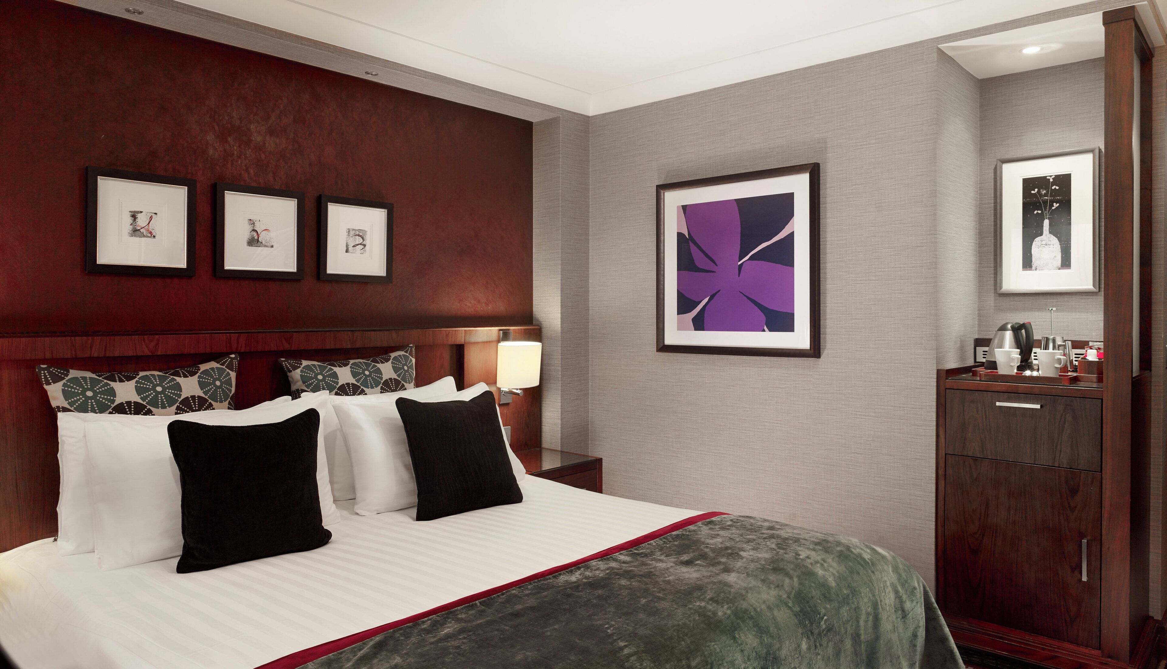 A hotel room with a large bed, a nightstand with a lamp, a dresser, and framed artwork on the walls.