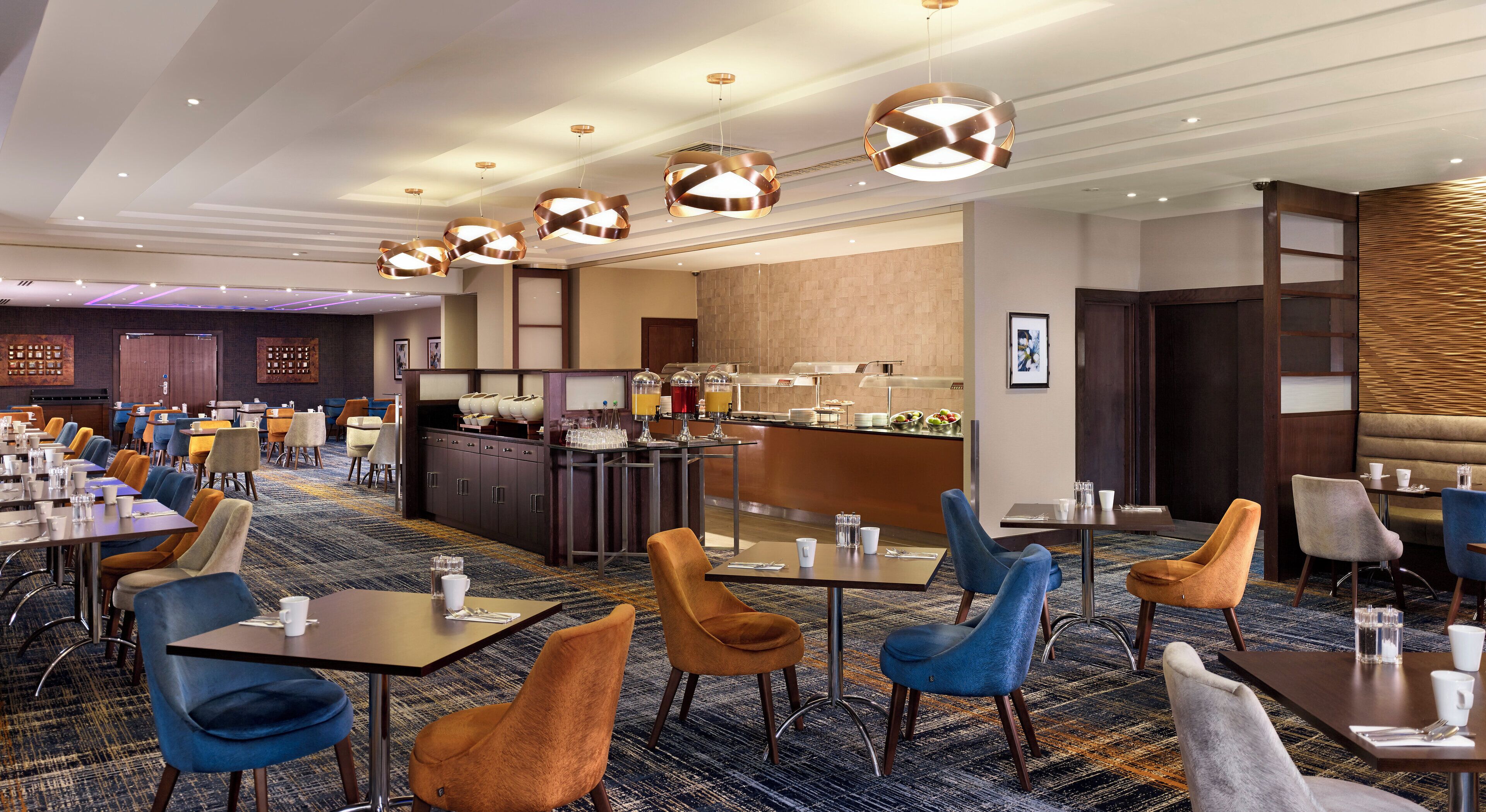 A hotel dining area with multiple tables and chairs, a buffet counter, and modern pendant lighting.
