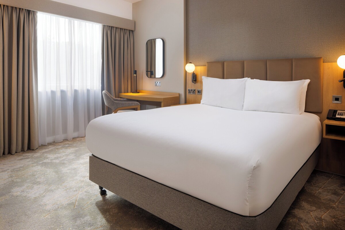Ein Hotelzimmer mit einem großen Bett, einem Schreibtisch, einem Sessel, einem Spiegel und einem Nachttisch mit einer Lampe.
