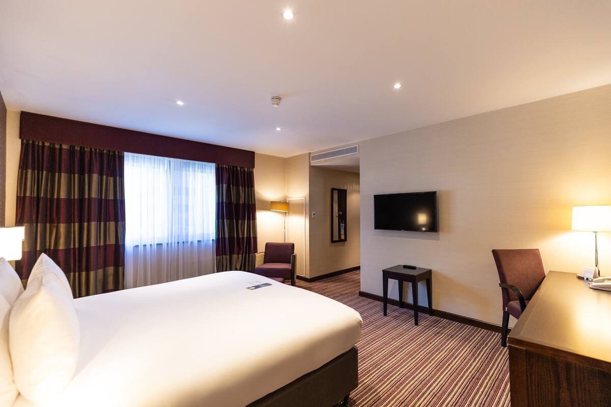 A hotel room with a large bed, a desk, a chair, a TV, and a window with curtains.