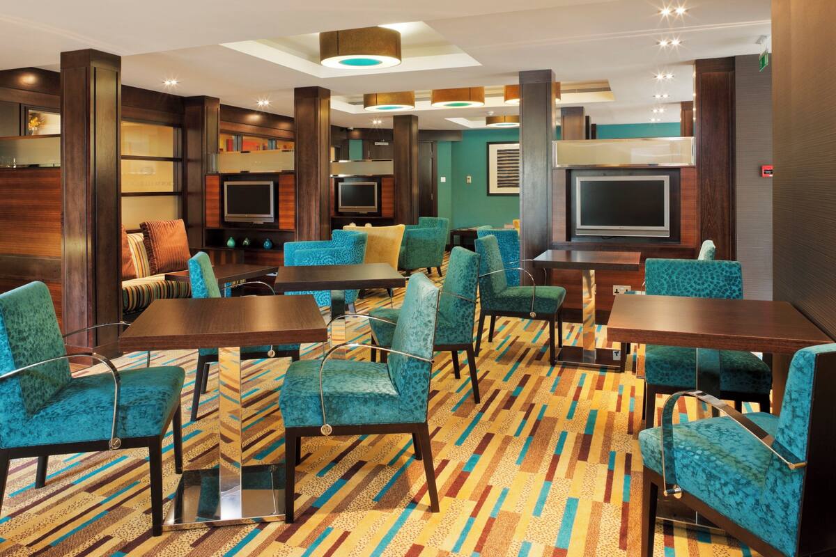 A hotel lobby with a patterned carpet, teal upholstered chairs, a wooden reception desk, and a television mounted on the wall.