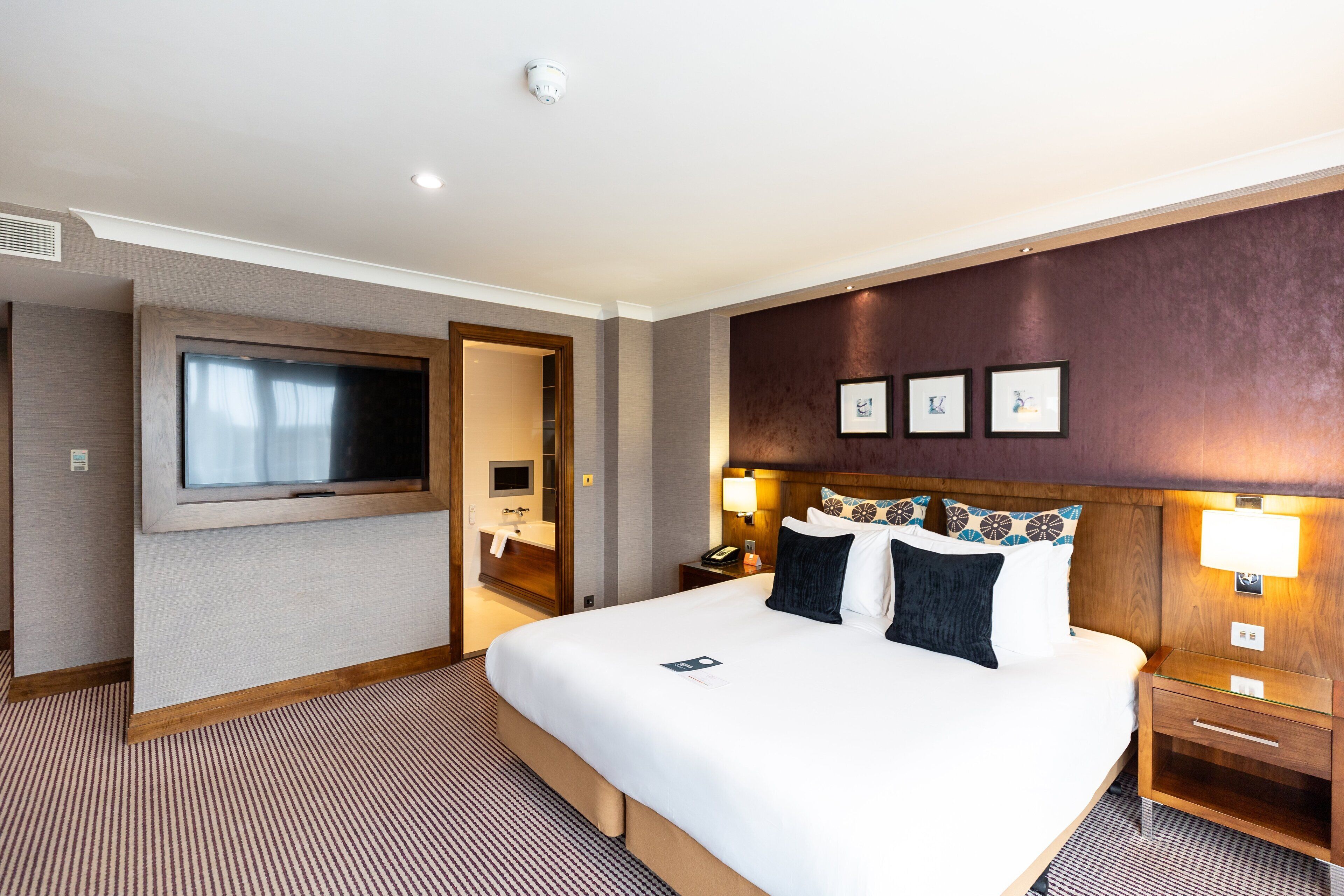 A hotel room with a large bed, a flat-screen TV, a bedside table with a lamp, and a wall-mounted picture.