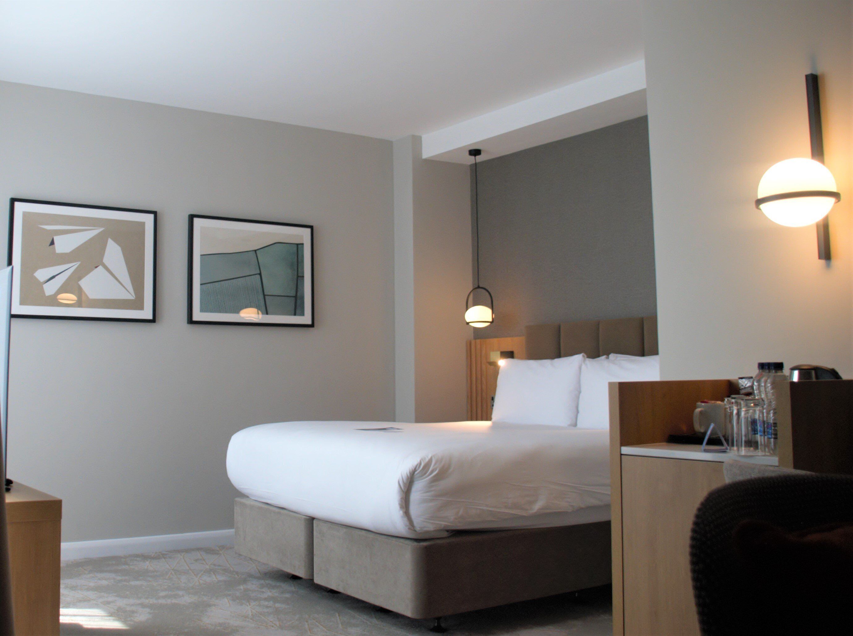 A hotel room with a bed, bedside table, lamp, and two framed artworks on the wall.