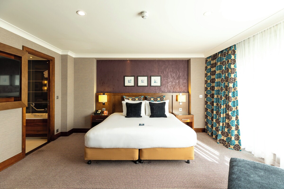 Habitación de hotel con una cama grande, una puerta de madera, una mesita de noche, una lámpara y una cortina estampada.