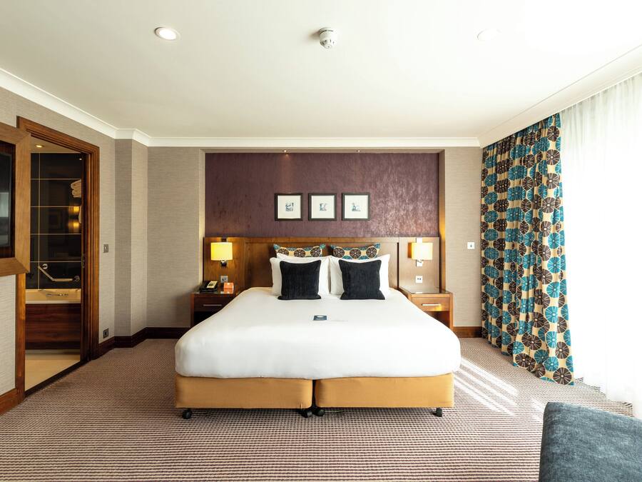 A hotel room with a large bed, a wooden headboard, a flat-screen TV, a desk with a lamp, and a patterned curtain.