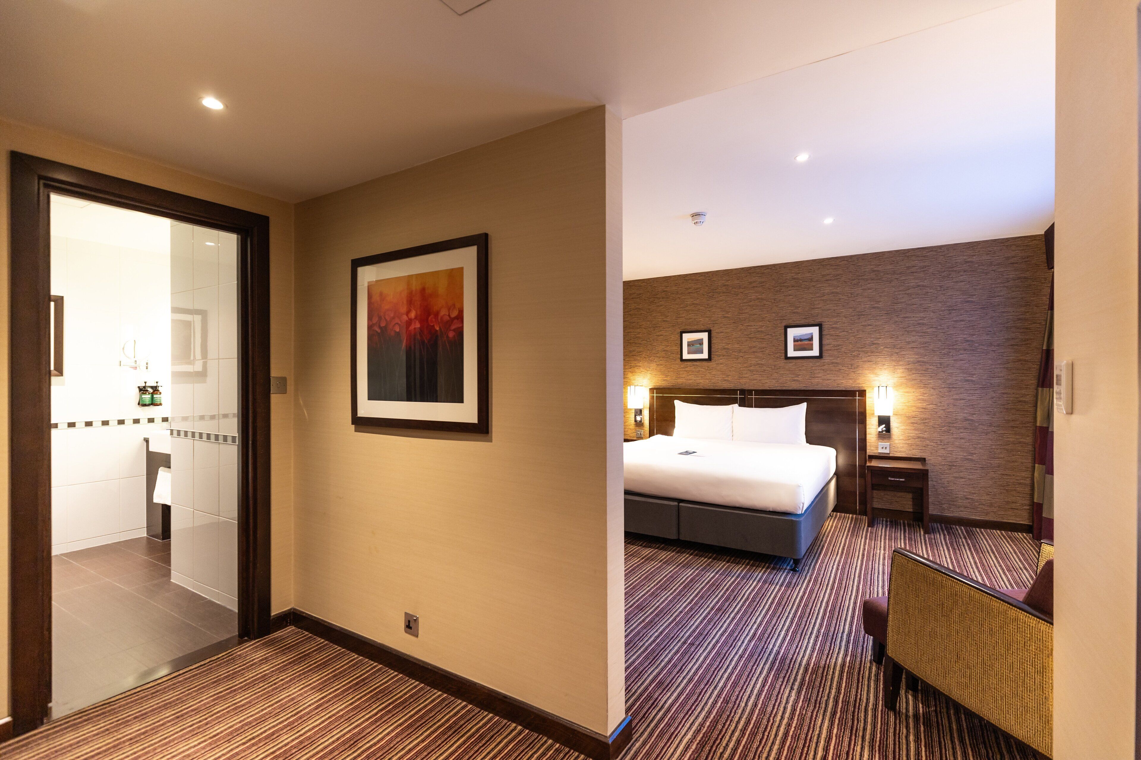 Een hotelkamer met een bed, een nachtkastje, een lamp, een foto aan de muur en een zichtbare badkamer door een open deur.