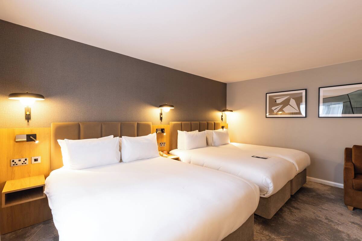 A hotel room with two beds, a wooden nightstand, wall-mounted lamps, and two framed pictures on the wall.