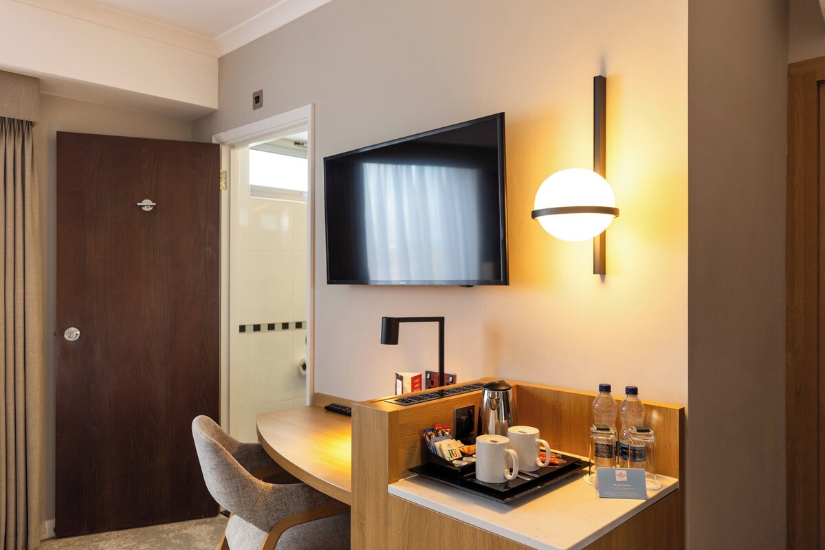 Habitación de hotel con un escritorio de madera, un televisor de pantalla plana montado en la pared, una cafetera y una bandeja con tazas y agua embotellada.