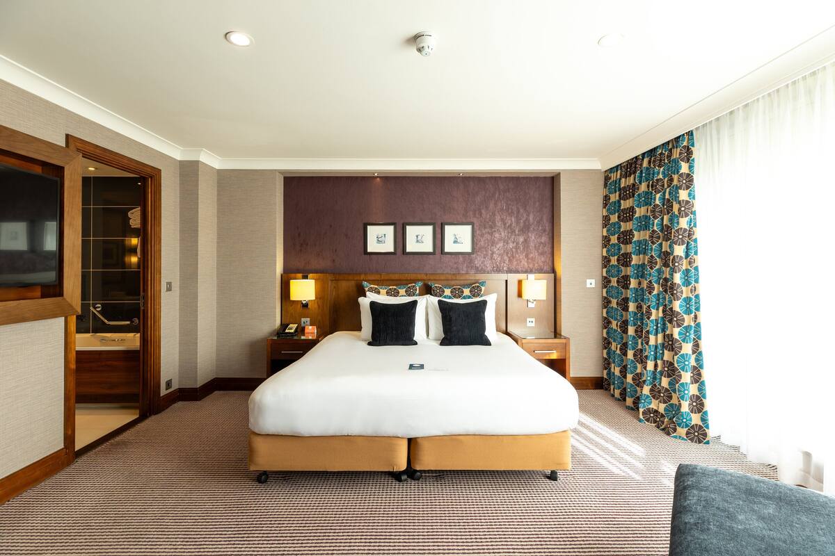 A hotel room with a large bed, a wooden door, a nightstand, a lamp, and a patterned curtain.
