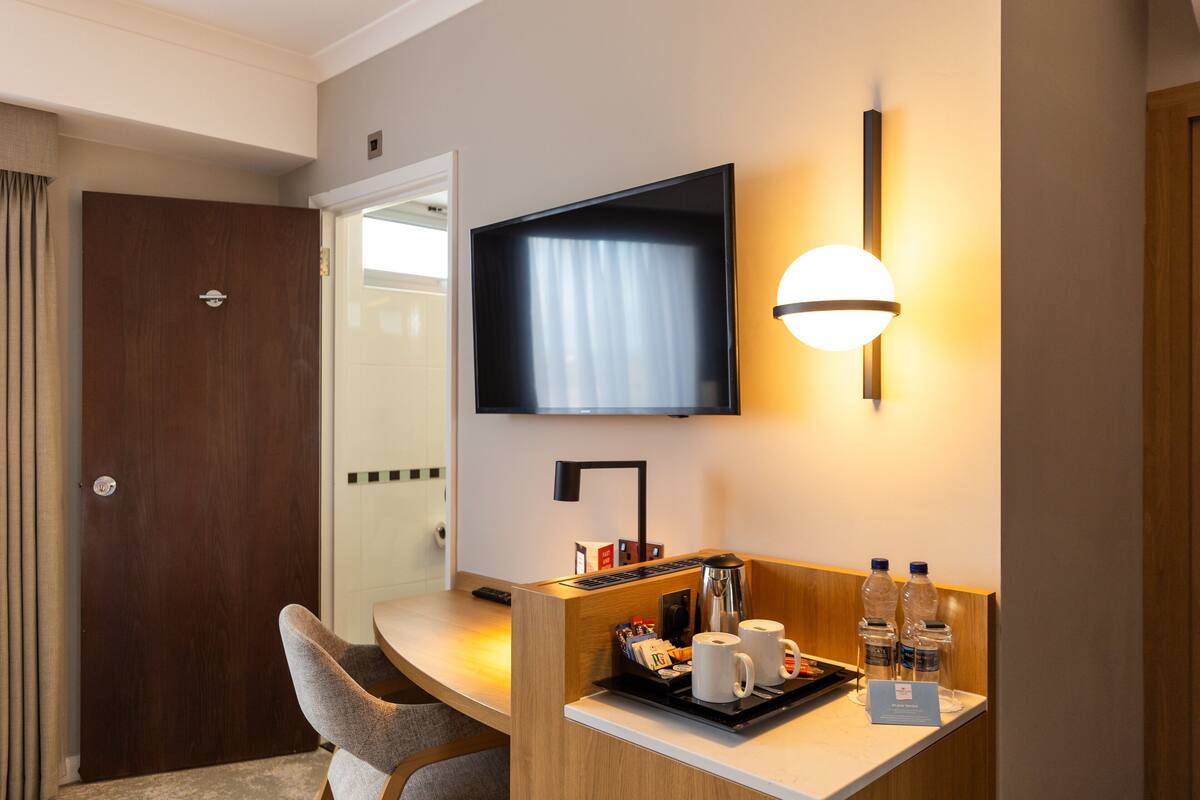 A hotel room with a wooden desk, a flat-screen TV mounted on the wall, a coffee maker, and a tray with cups and bottled water.