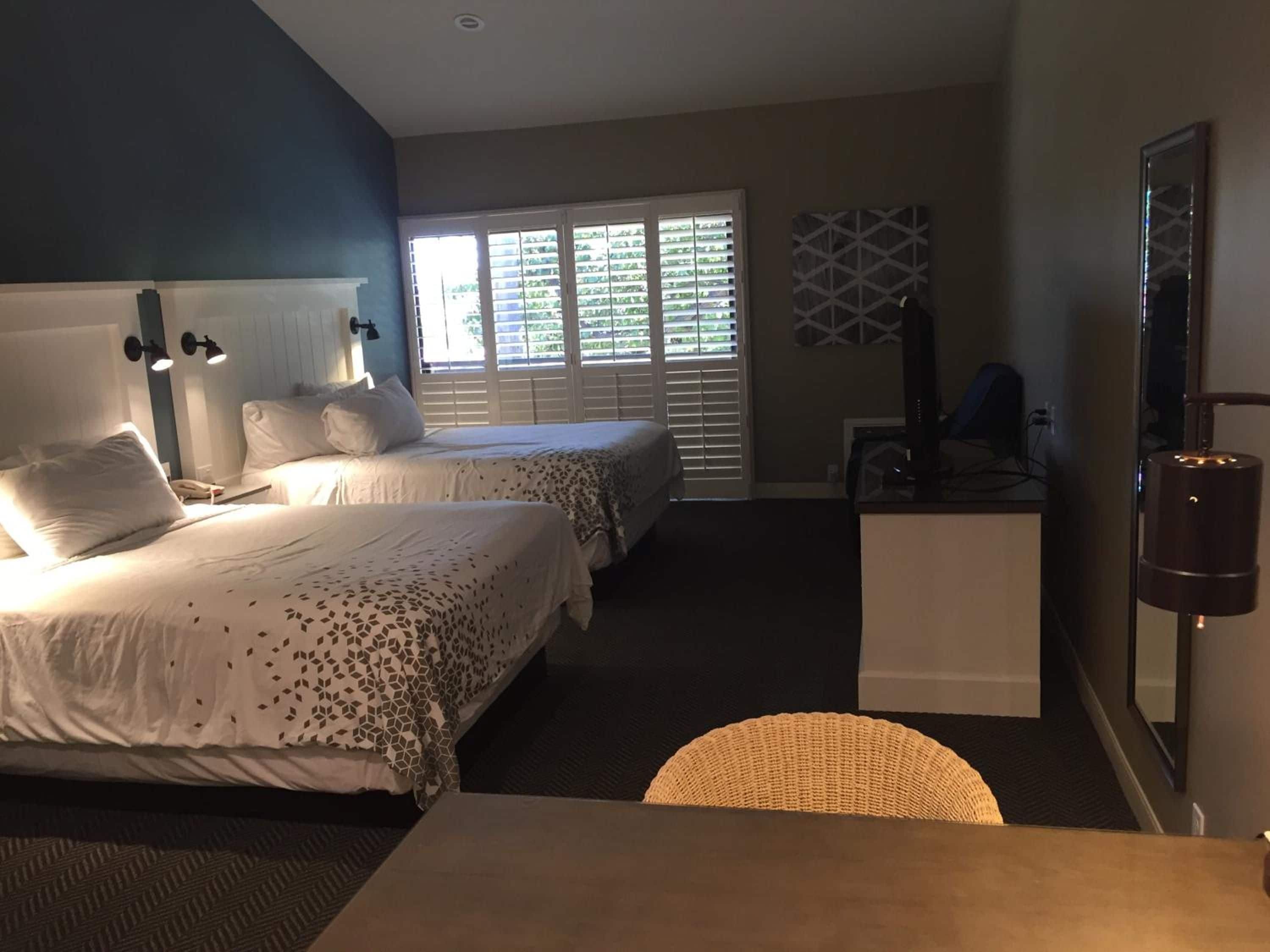 A hotel room with two beds, a desk, a chair, a TV, and a window with blinds.