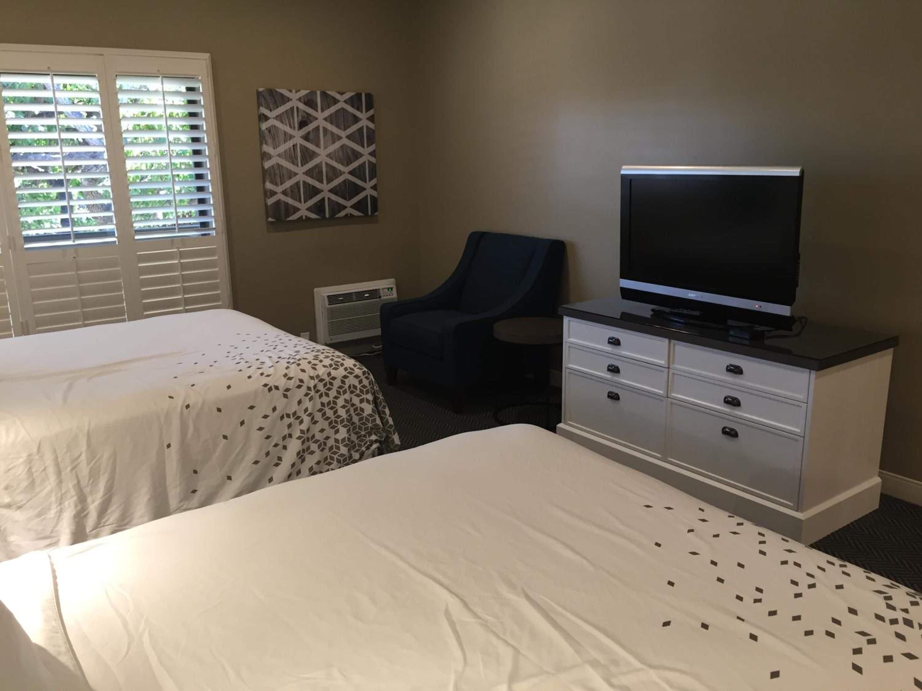 A hotel room with two beds, a television on a dresser, and a window with blinds.