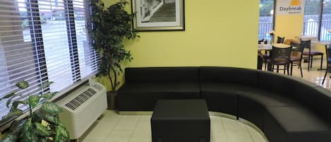 Un área de recepción con un sofá negro curvo, una planta en maceta, una fotografía enmarcadas en la pared y un cartel que anuncia 'Daybreak Breakfast'.