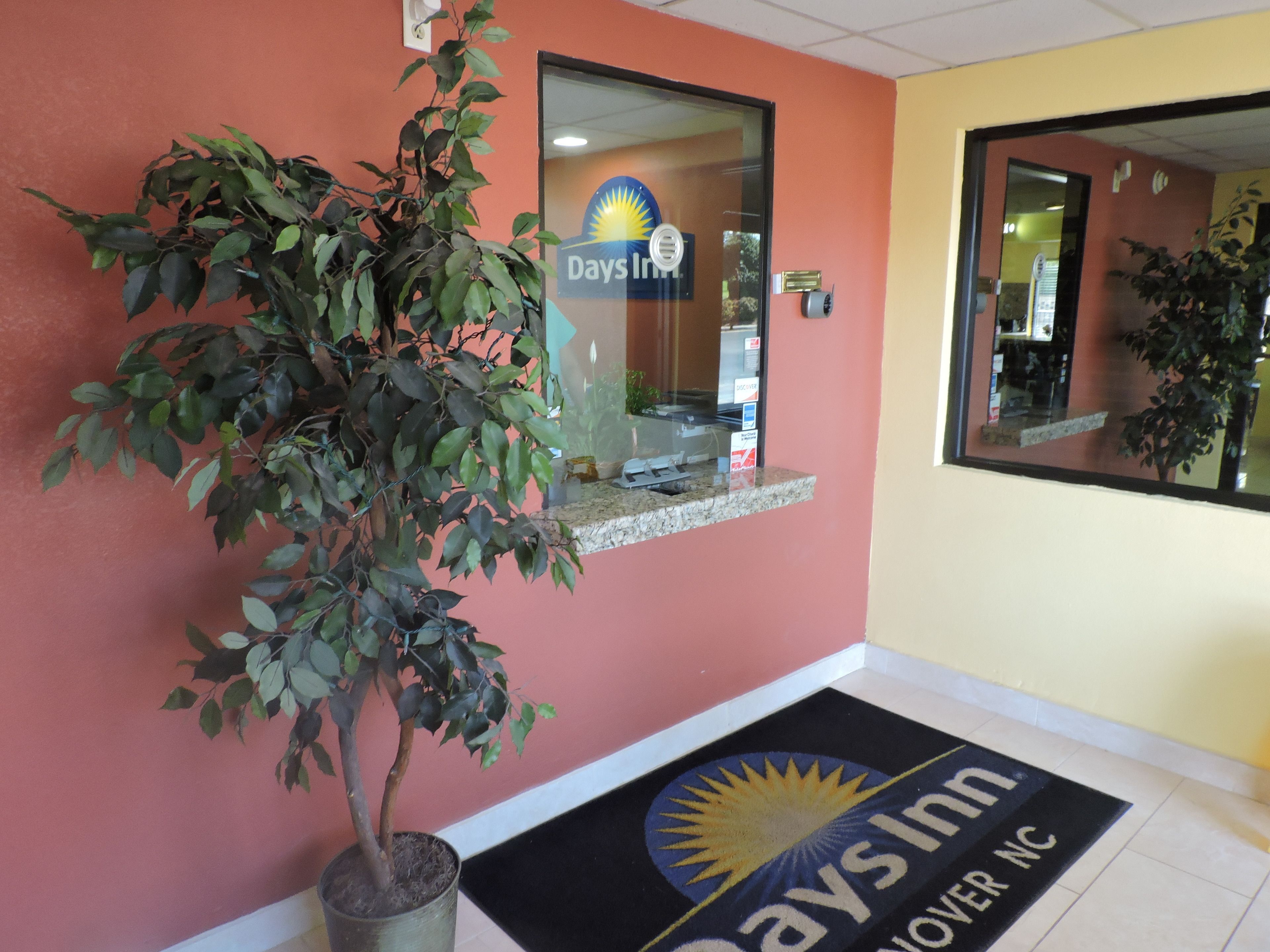 A reception area with a potted plant, a reception desk, and a doormat with the hotel's name.