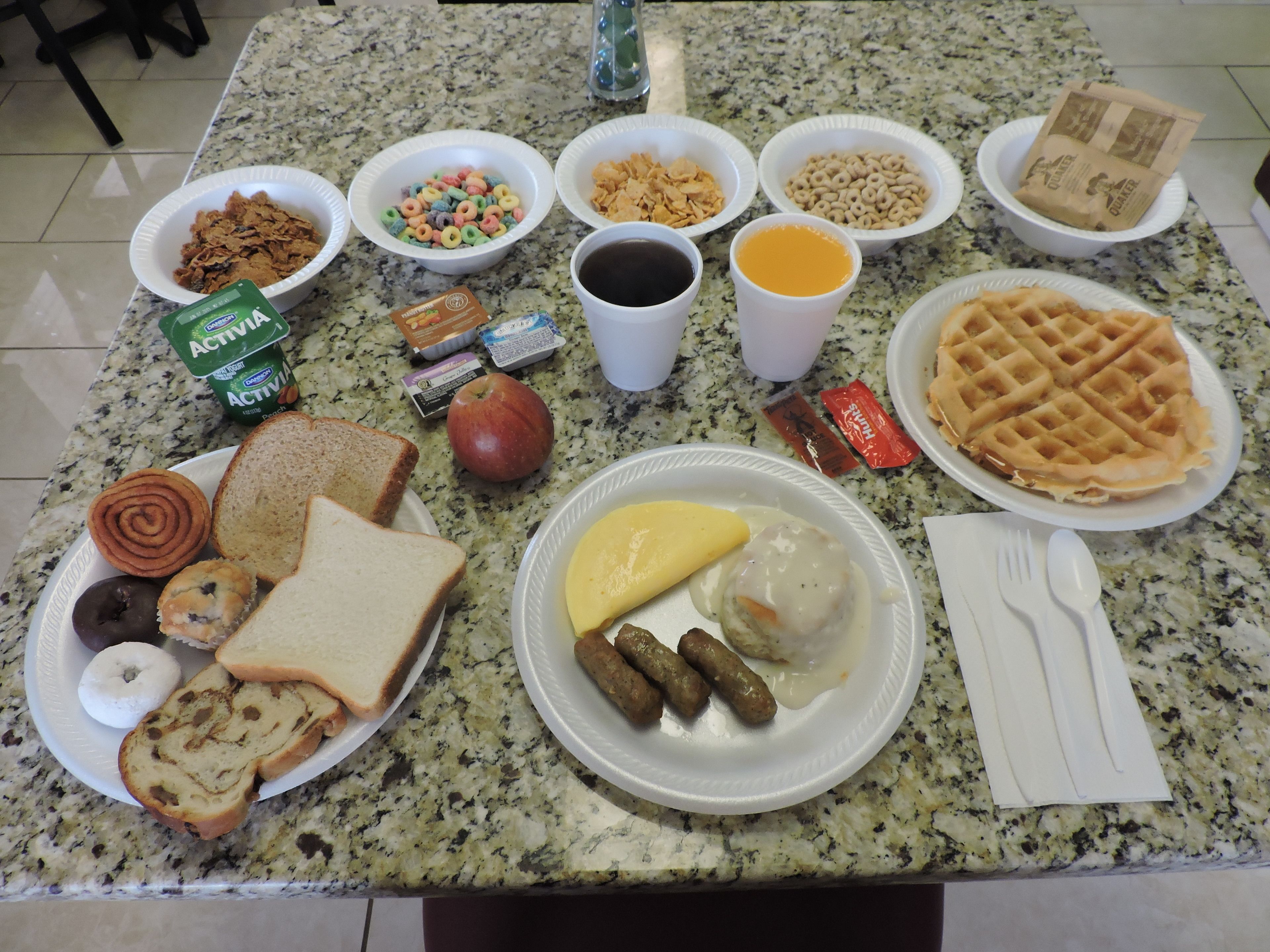 A breakfast spread on a granite countertop including cereal, toast, waffles, sausages, and beverages.
