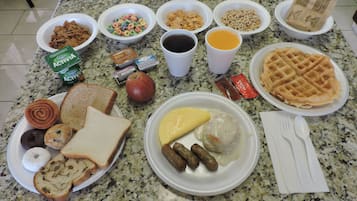 A breakfast spread on a granite countertop including cereal, toast, waffles, sausages, and beverages.