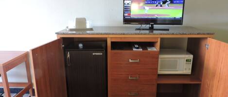 A compact kitchenette with a refrigerator, microwave, and a television displaying a baseball game.