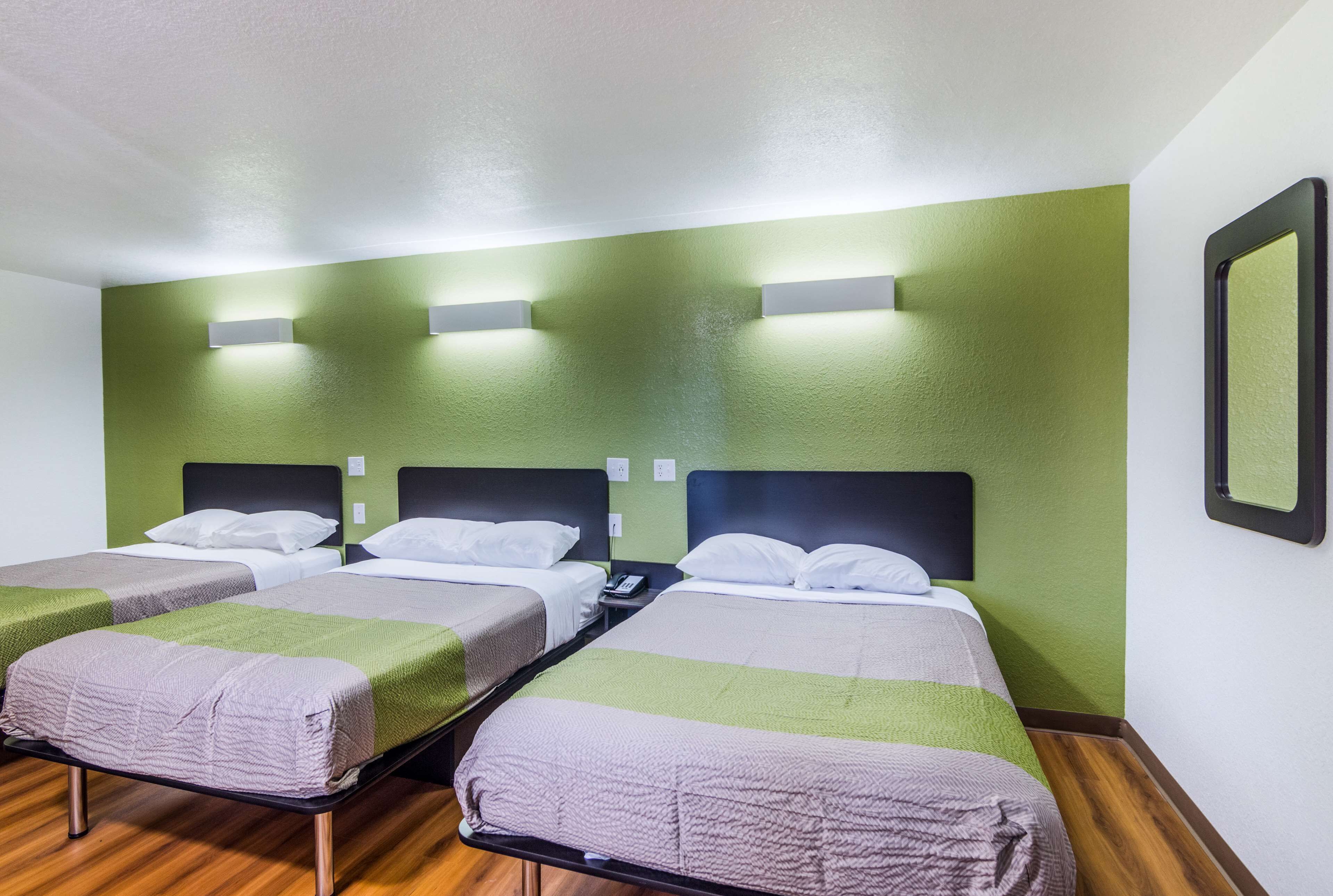 A hotel room with three single beds, each with a green and white striped blanket. There are wall-mounted lights above each bed and a mirror on the wall.
