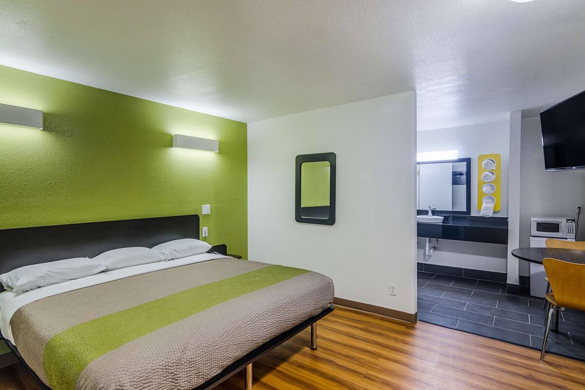Ett hotellrum med en säng, ett litet bord, en stol, en spegel, en TV och ett litet kök.