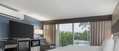 A hotel room with a large bed, a desk with a computer, a TV, and a view of trees through large windows.