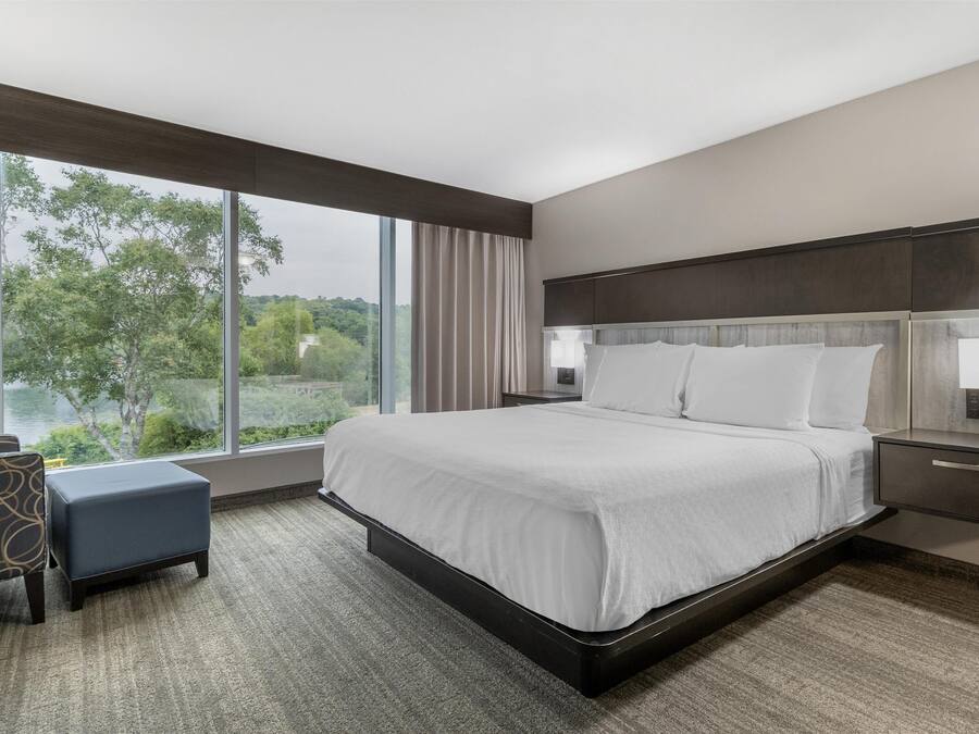 A modern hotel room with a large bed, a chair, and a view of trees through a window.