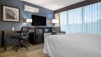 Habitación de hotel con cama, escritorio, silla, televisor, equipo de aire acondicionado y ventanas grandes.