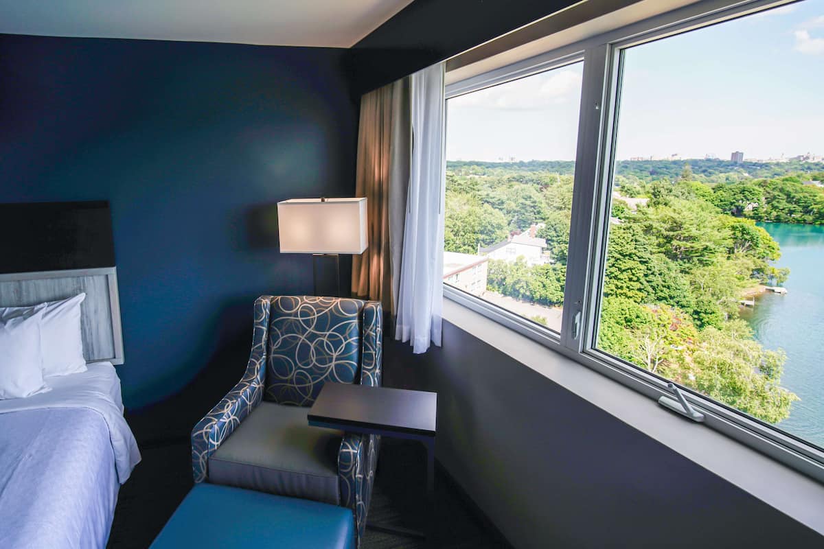 A hotel room with a bed, a chair, a small table, a lamp, and a large window offering a view of a lake and greenery.