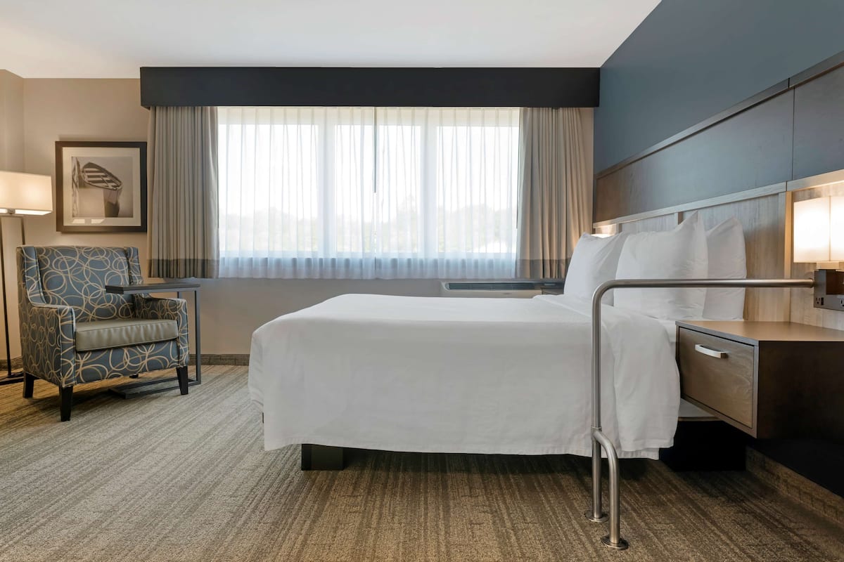 A hotel room with a bed, bedside table, lamp, chair, and a large window with curtains.