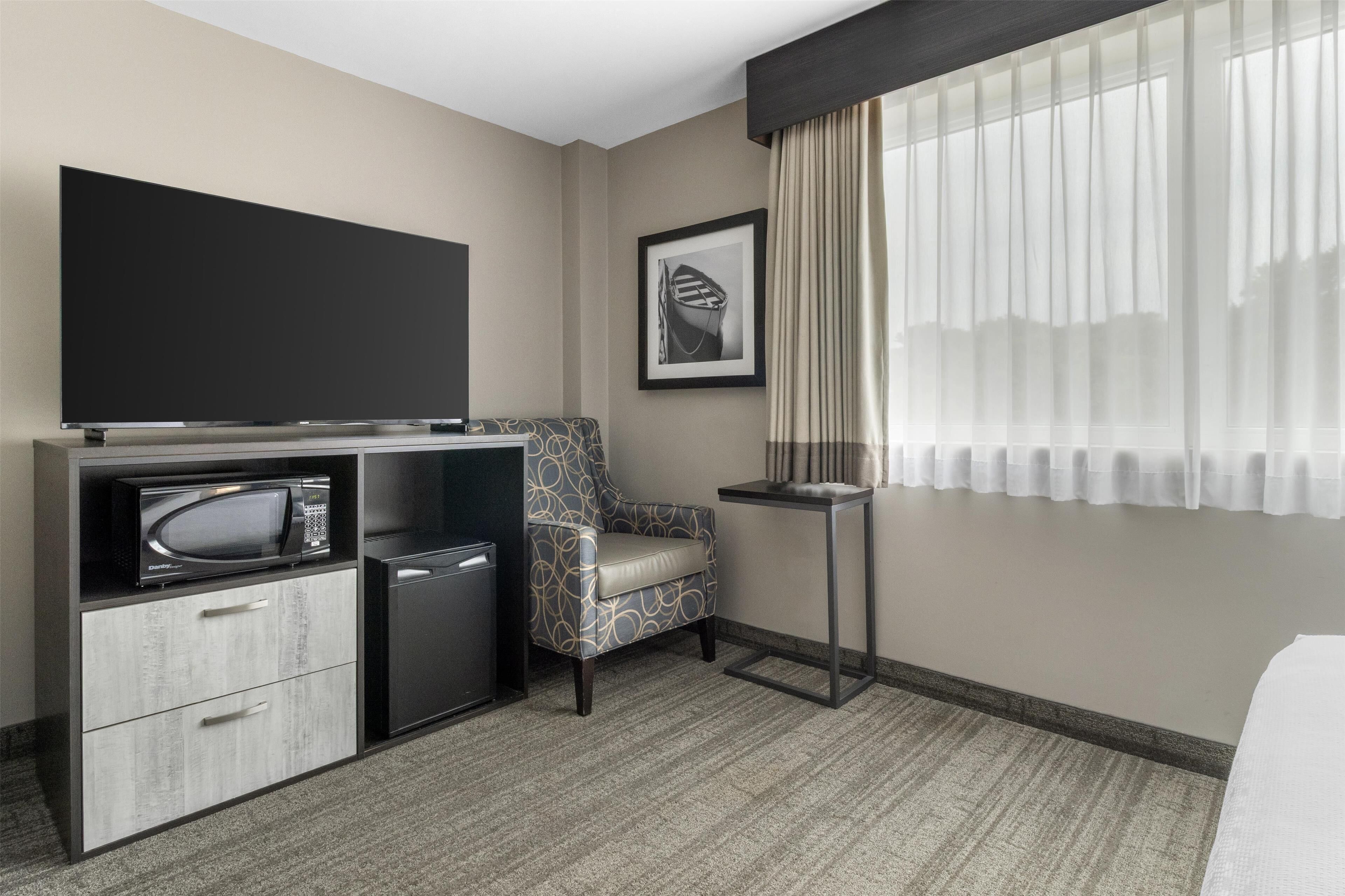 Habitación de hotel con televisión de pantalla plana, microondas, refrigerador pequeño, sillón estampado, una mesita y un ventanal grande con cortinas translúcidas.