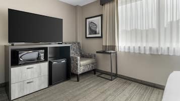 Une chambre d’hôtel équipée d’un téléviseur à écran plat, d’un four à micro-ondes, d’un mini-réfrigérateur, d’un fauteuil à motifs, d’une petite table et d’une grande fenêtre avec des rideaux légers.