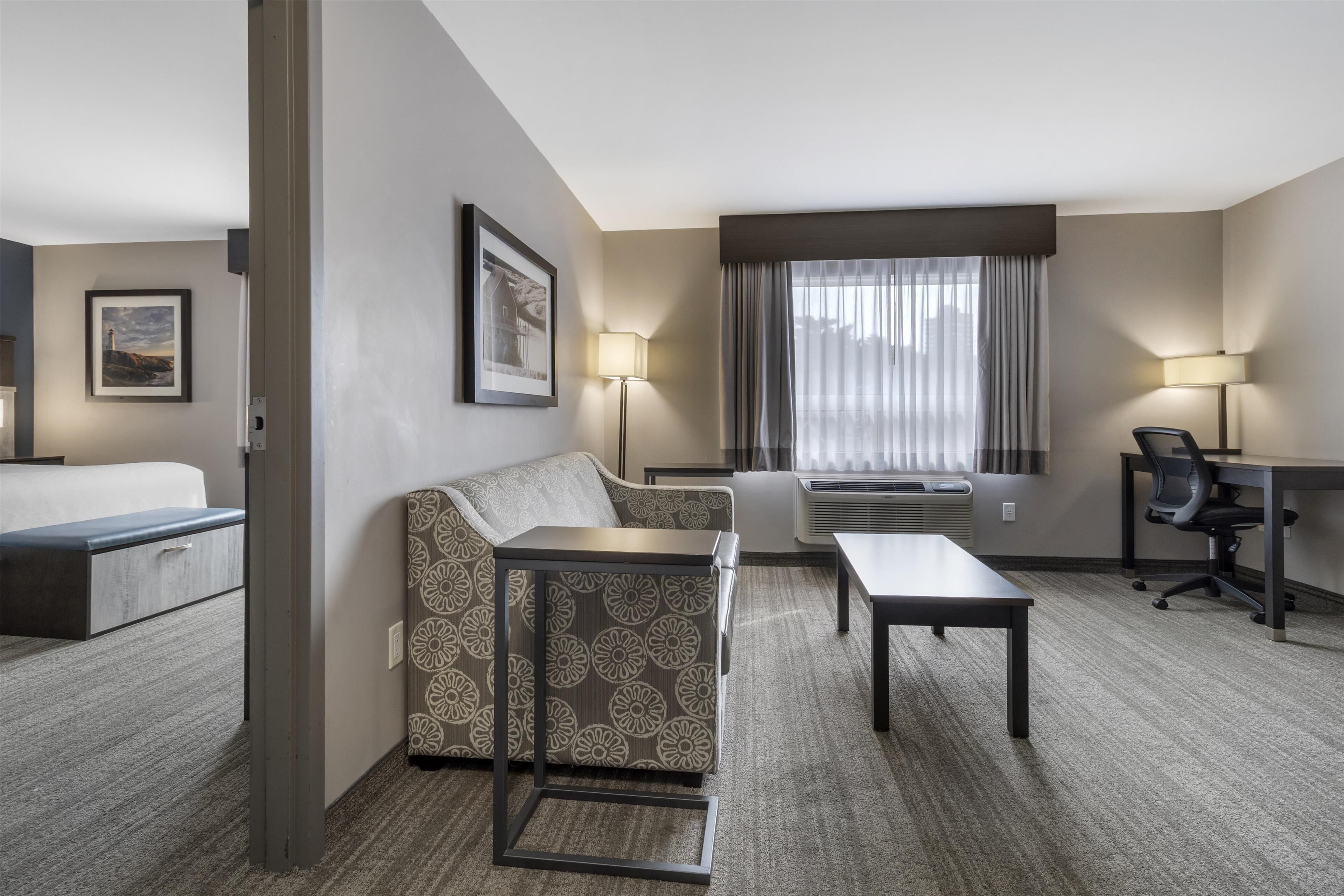 A hotel room with a bed, a sofa, a small table, a desk, and a chair. There are two lamps and a window with curtains.