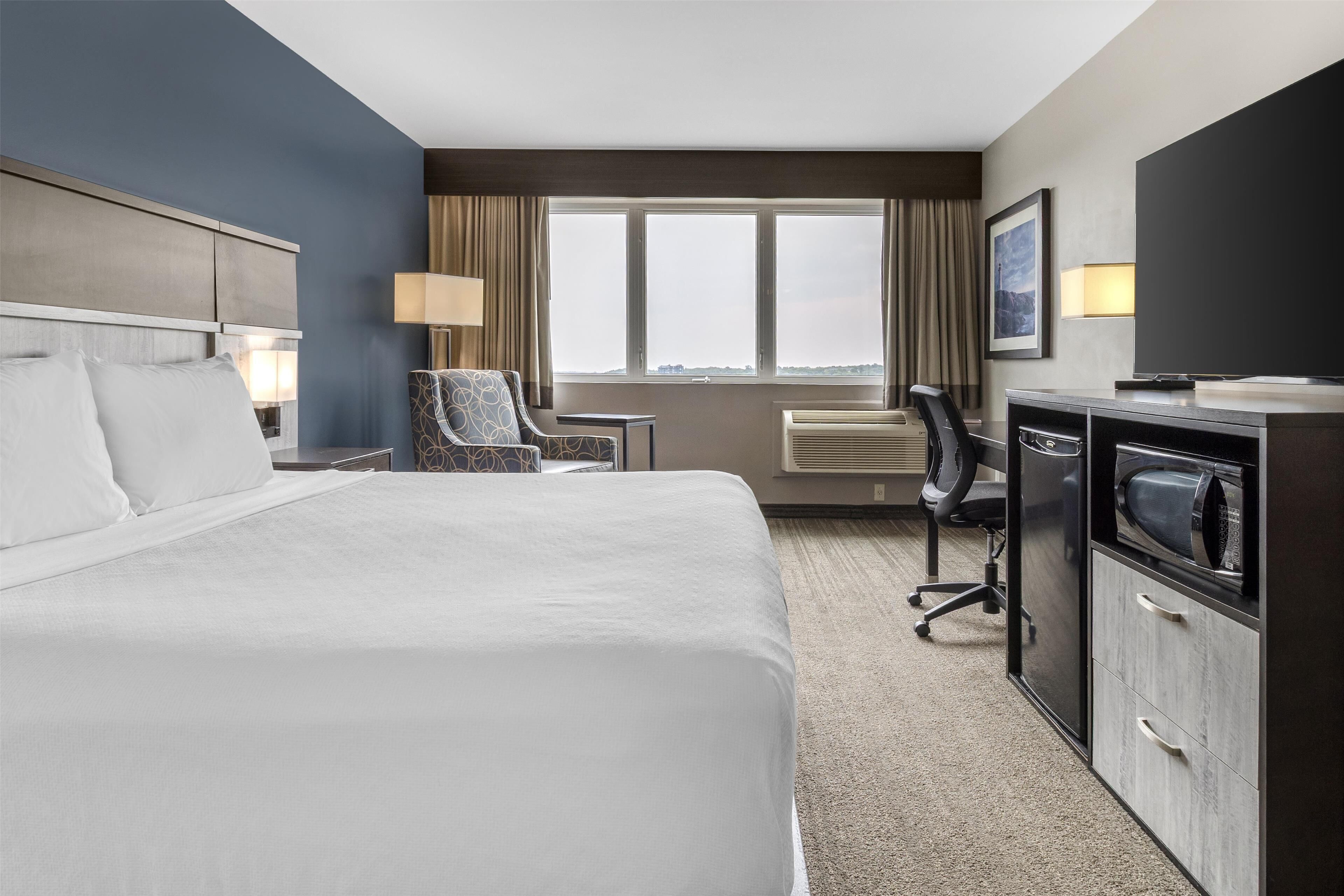 Ein Hotelzimmer mit einem großen Bett, einem Schreibtisch mit Computer, einem Sessel, einem Fernseher und einem Fenster mit Vorhängen.
