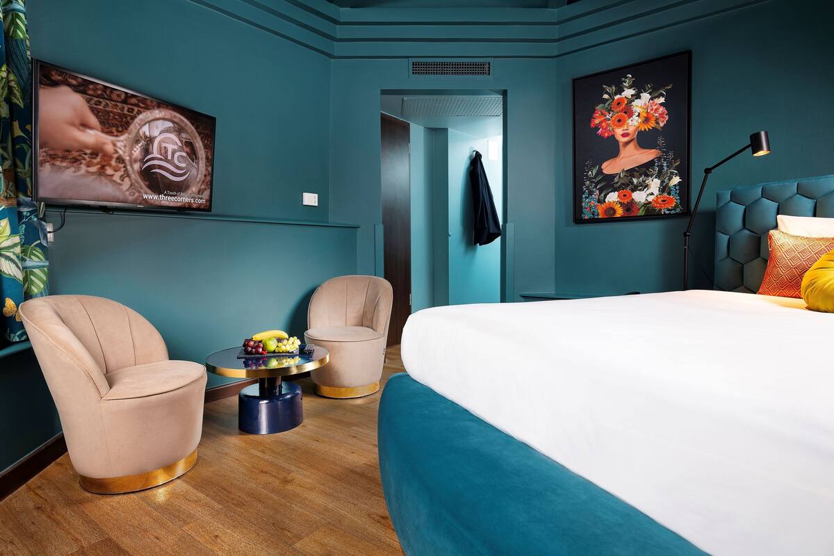 A modern hotel room with a large bed, two armchairs, a small round table with fruit, a painting on the wall, and a TV displaying a website.