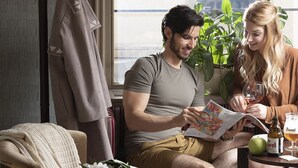 A couple sitting on a couch reading a book, with a window and a plant in the background. - Three Corners Hotel Art (Budapest)