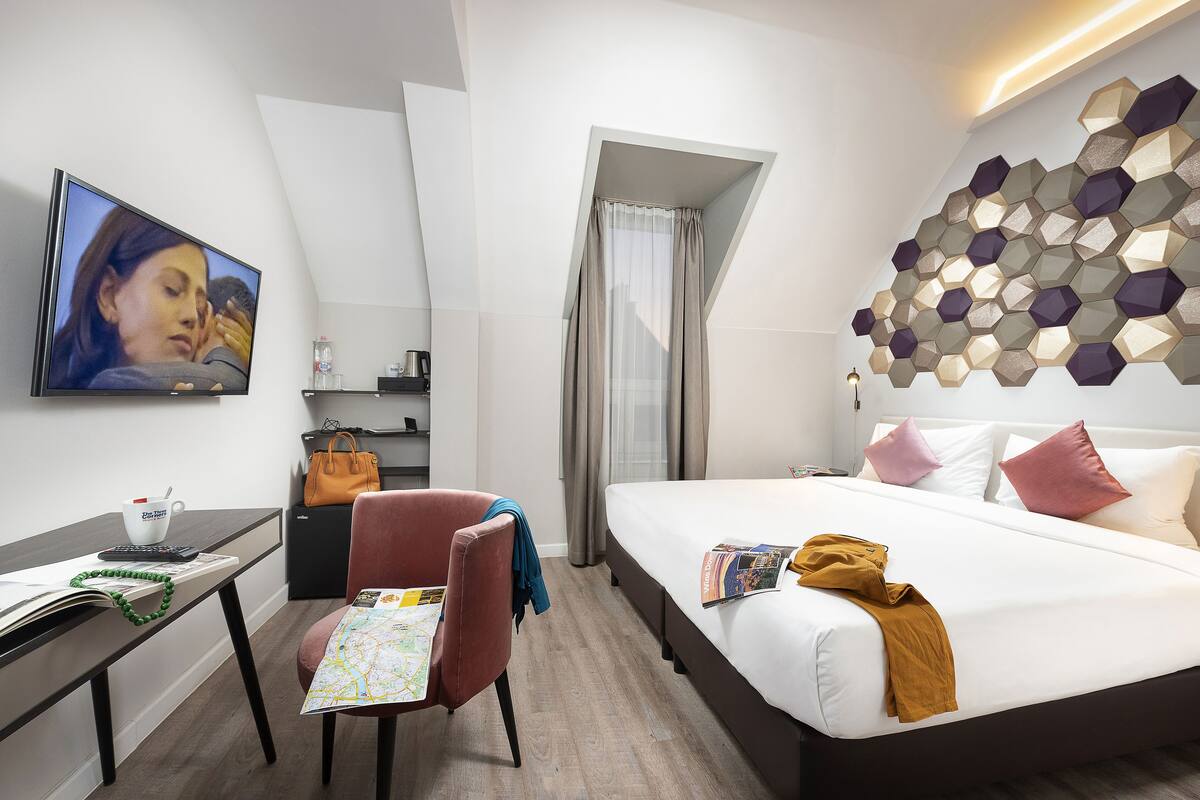 A modern hotel room with a bed, a desk, a chair, a TV, and a unique hexagonal wall design.