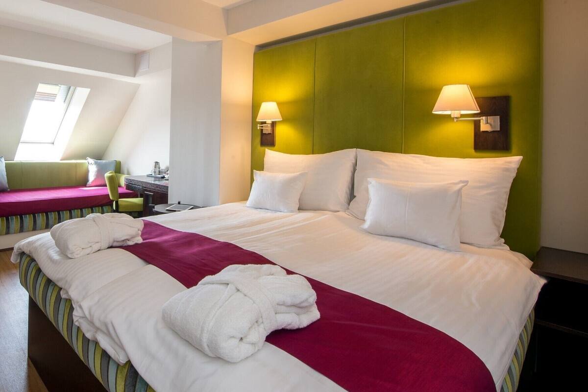 A hotel room with a large bed, two bedside lamps, a green headboard, and a view of the sky through a skylight.
