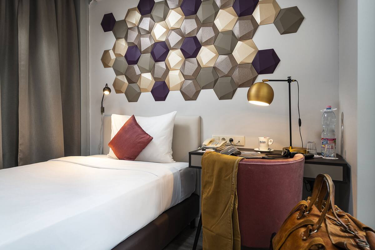 A hotel room with a neatly made bed, a wall-mounted phone, a bedside table with a lamp, a water bottle, and a handbag.